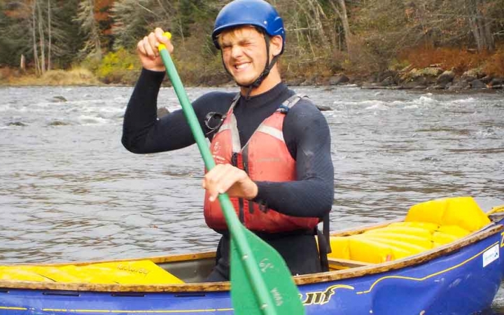 a person wearing a life jacket and helmet smiles while paddling a canoe on an outward bound expedition 